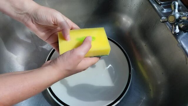 Unas manos de  mujer lavando un plato  de color blanco  con una esponja, jab&oacute;n liquido y  agua  en un fregadero