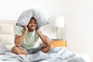 Unhappy black man sitting on bed, covering head with pillow
