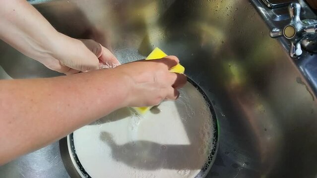 Unas manos de  mujer lavando un plato de color blanco  con una esponja con jab&oacute;n liquido  en un fregadero
