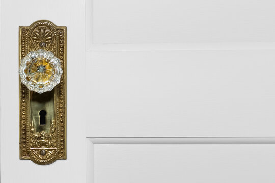 Close-up side view photo of a beautiful crystal door handle on a brightly polished ornate gold plate with a skeleton keyhole. White raised panel wooden door background with copy space.