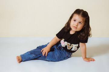 Hermosa niña blanca de dos años de cabello ondulado, sentada en el suelo pensativa, con camiseta...