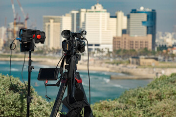 A professional video camera and video light stand on a tripod, filming a general shot against the backdrop of the sea and the city.