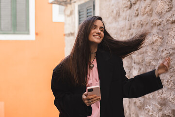 Photo of charming pretty young lady hold modern gadget smiling outside urban city street. Pensive brunette woman student freelancer wear black jacket walking in old town, hold phone in hand.