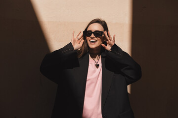 Portrait of brunette attractive woman puts on sunglasses and smiles widely near beige wall with sun...