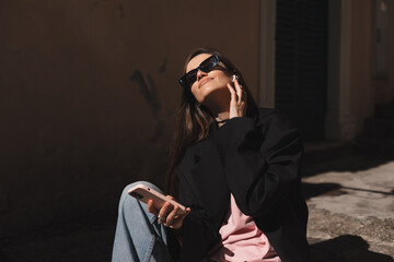 Smiling young woman sitting on stairs and touch wireless earphones. Woman is relaxing listening music in old town. Young woman using technologies, wear black glasses, black jacket, hold mobile phone.