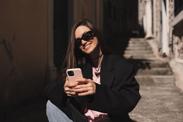 Young stylish brunette woman writes message on smartphone walking on old town street, hipster tourist traveling and planning trip using mobile phone. Look happy when read message, sitting on stairs.