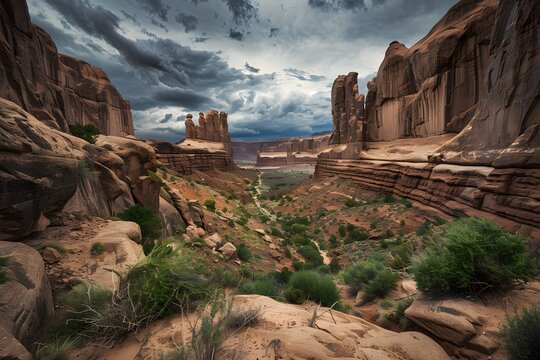 Expansive view of Arches National Park: towering canyons, narrow trails, and green shrubs against a dramatic sky over brown sandstone cliffs.