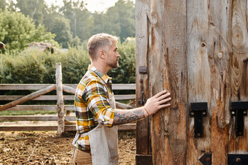attractive dedicated man with beard and tattoos on his arms closing barn doors while on farm