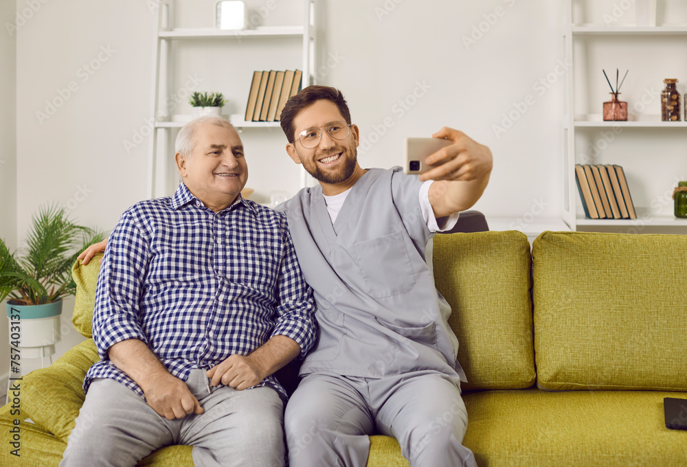 Wall mural Senior man and young caregiver taking selfie photo at home. Smiling male social worker and senior patient sitting on couch making photo and smiling at camera. Elderly people care and support