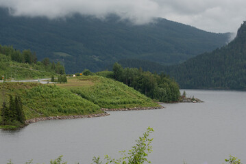 river in Norway flowing along mountains with green trees and next to a highway on a foggy day.