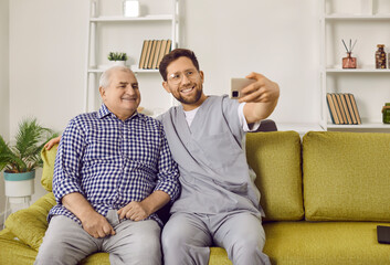 Senior man and young caregiver taking selfie photo at home. Smiling male social worker and senior patient sitting on couch making photo and smiling at camera. Elderly people care and support