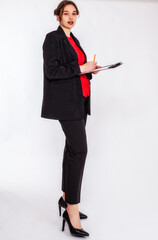Eine junge, blonde Frau mit Dutt trägt einen schwarzen Hosenanzug und eine rote Bluse. Sie hält ein Schreibbrett und einen Bleistift in der Hand und schaut konzentriert in die Kamera. 