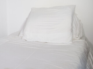 Closeup of white pillows on a single bed, covered with a white bed quilt, against plain white room walls.