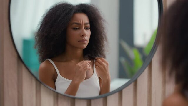 Worried beautiful sad upset African American woman girl female lady looking at mirror reflection holding fragile curly hair damaged dry curls unhealthy split ends hormone problem vitamins deficiency
