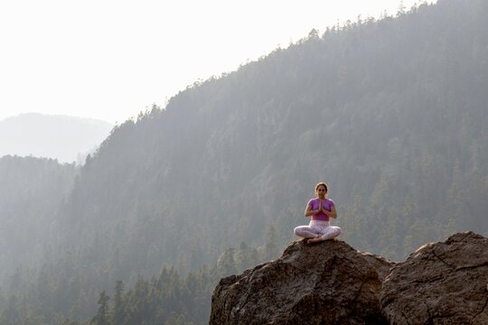 A person sits in lotus pose, meditating on a mountain top with a hazy forested background, evoking serenity and mindfulness