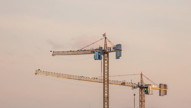 Large construction site including several cranes working on a building complex, with golden sky during sunset timelapse. Clouds passing by