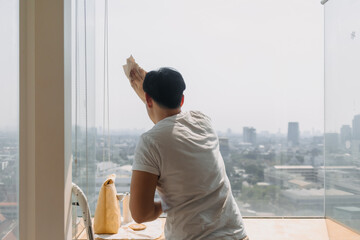Backside view of asian Thai man wiping window glass in room apartment with city view, keep glass...