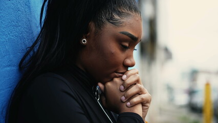 One pensive young black woman pondering decision with hands clenched together standing outside with thoughtful gaze struggling with dilemma in in urban setting - obrazy, fototapety, plakaty