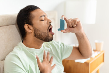 Handsome black man sitting alone at home, using asthma pump