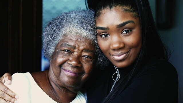 Happy portrait of young African American granddaughter with arm around her grandmother posing for camera, close-up faces of intergenerational family members embrace