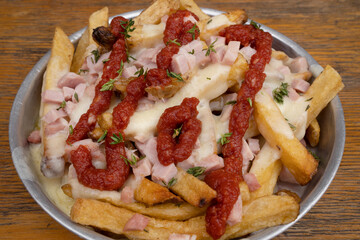 Fried potatoes with ham, mozzarella cheese, ketchup, onion and fresh oregano in a metal dish with a wooden background.