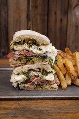 Closeup view of a slice din half gourmet sandwich with layers of ciabatta bread, prosciutto ham, pesto sauce, burrata cheese and tomato, with french fries and a rustic wooden background
