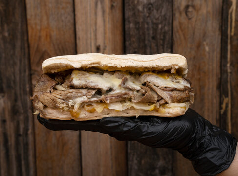 Closeup view of the chef hands wearing black gloves holding a meat sandwich made with ciabatta bread, meat, mozzarella cheese, chucrut and mustard with french fries and a rustic wooden background.
