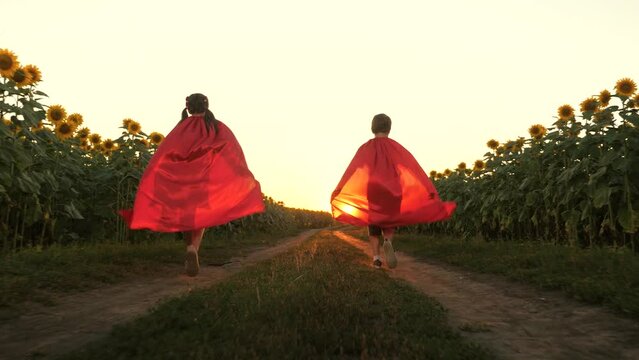 Boy and girl superhero in red cloak running flying on road at sunflower field sunset back view. Happy kids playing fantasy imagination pretend powerful hero character planet protect enjoy childhood