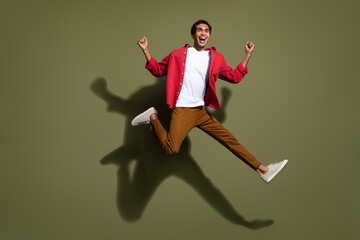 Fototapeta na wymiar Full length photo of positive lucky man dressed red shirt jumping high rising fists empty space isolated green color background