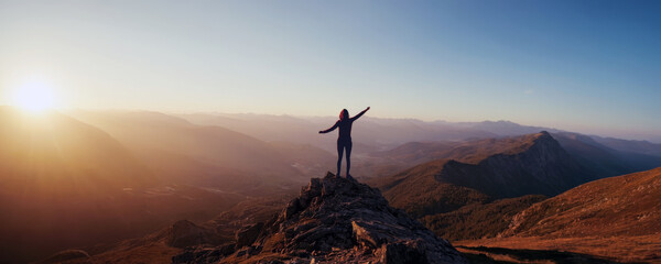 A woman opens her arms as she looks down from a mountain peak at several other mountains and...