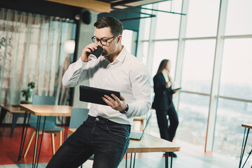 A young man in a white shirt stands in a spacious modern office with coffee and works on a tablet....