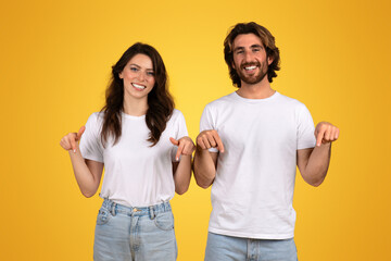 Smiling woman and man in white shirts pointing down with both hands, appearing cheerful and confident