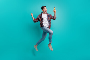 Fototapeta na wymiar Full length photo of cheerful lucky guy wear checkered shirt jumping high winning game modern device isolated teal color background