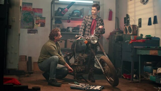 Maintenance service and small family business concept. Professional repairman and young boy working in home garage. Mechanic servicing or repairing vintage retro motorbike with son in workshop.