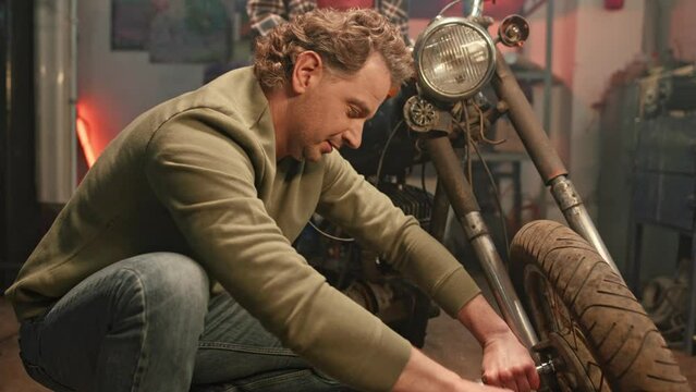 Mechanic servicing or repairing vintage retro motorbike in neat well equipped workshop. Professional repairman looking at camera and smiling while working in garage indoors. Maintenance service.