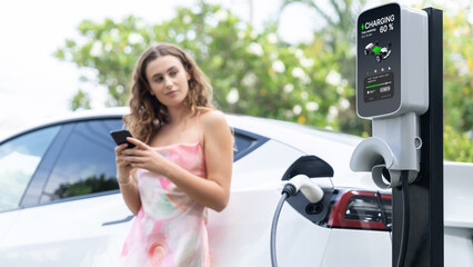 Focused charging station recharging electric vehicle on blurred background of modern woman using...
