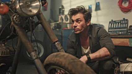 Portrait of male mechanic spinning wheel of retro motorcycle. Young biker tuning motorcycle in workshop. Auto mechanic inspecting vintage motorbike condition in garage. Biker lifestyle concept. - Powered by Adobe