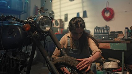 Beautiful female mechanic inspecting vintage motorbike condition in garage. Young woman biker tuning motorcycle in workshop. Retro motorcycle parts. Girl biker lifestyle concept. - Powered by Adobe