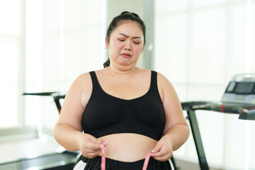 Plus-size Asian woman exercise in gym, visibly upset while measuring waist, moment of struggle on fitness journey, Emotionally overwhelmed with measuring tape, confronting weight loss challenges