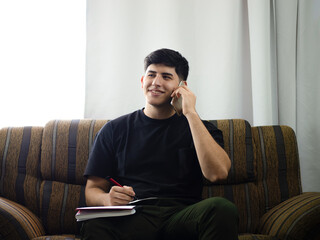 Image of a smiling young man talking cheerfully on the phone, sitting on his couch at home and taking notes of data passed to him during the call