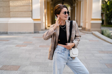 Fototapeta premium stylish woman with short haircut walking in street with coffee wearing beige jacket, jeans and sunglasses accessories