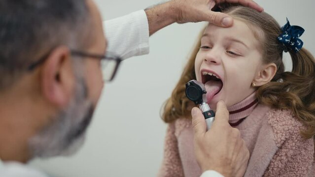 Shooting over the shoulder. A pediatrician performs a visual examination of the throat of a patient sitting on an examination couch in a modern clinic. A man doctor examines a little girl. The girl