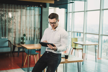 A young man in a white shirt stands in a spacious modern office and works on a tablet. Modern office worker. A businessman in glasses works online.