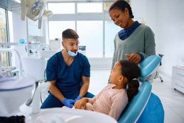 Young dentist talking to black little girl and her mother at dental clinic.