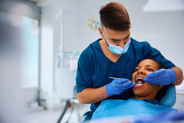 Male dentist examining teeth of black woman at dental clinic.