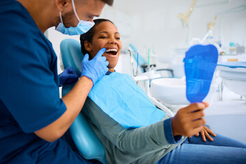 Black woman using mirror while examining her teeth with dentist at dental clinic.