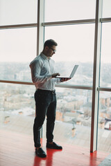 A young handsome man with a laptop stands on the background of a large panoramic window in the sky. A modern office with large windows and an office worker