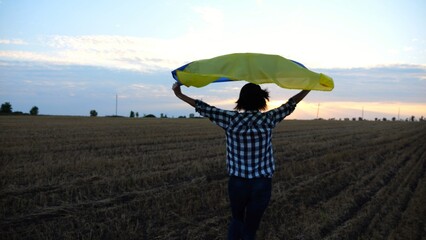 Ukrainian woman running with raised flag Ukraine above her head on wheat field at sunset. Lady...