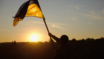 Young man in military uniform waving flag of Ukraine against beautiful sunset at background. Male ukrainian army soldier lifted national banner at countryside. Victory against russian aggression - 757492964