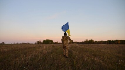 Soldier of ukrainian army running with raised blue-yellow banner on field at dusk. Young male military in uniform jogging with flag of Ukraine at meadow. Victory against russian aggression concept - 757493184